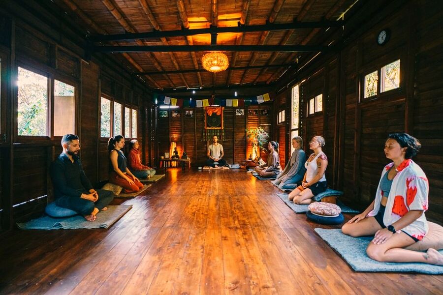 Wide view of a group meditation session in the meditation hall with prayer flags and the instructor facing the group